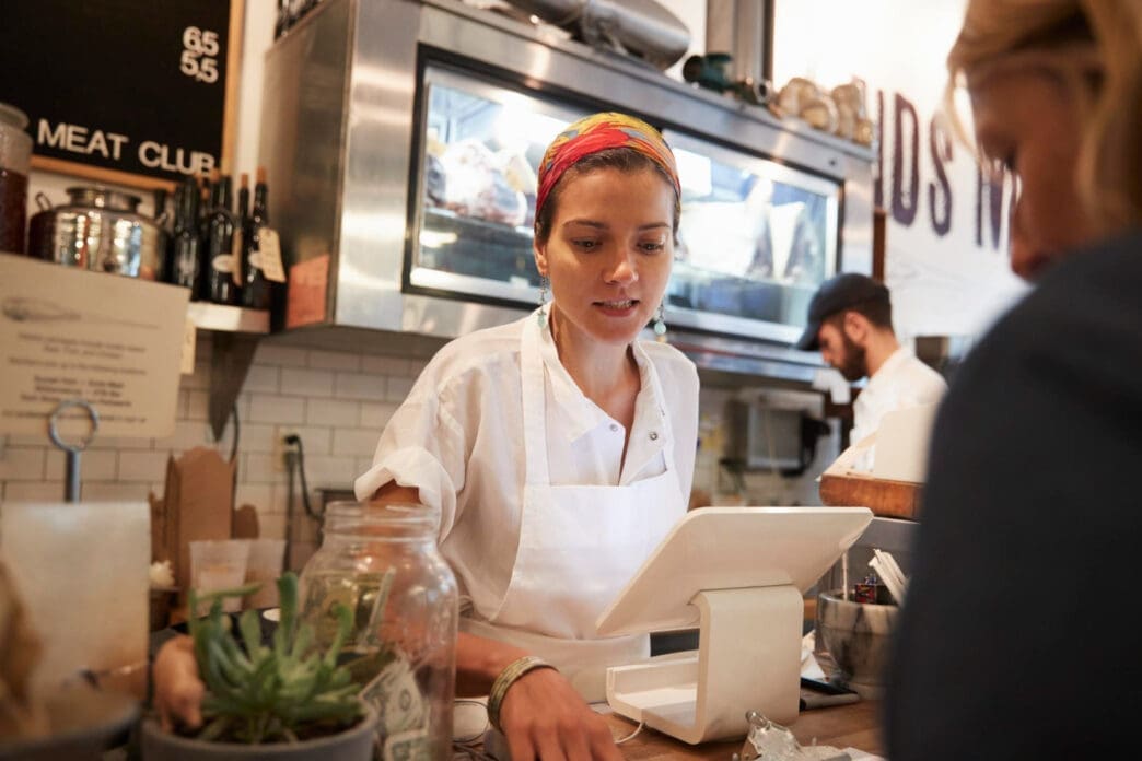 A small business owner in an apron and colorful headscarf works at the counter of her shop