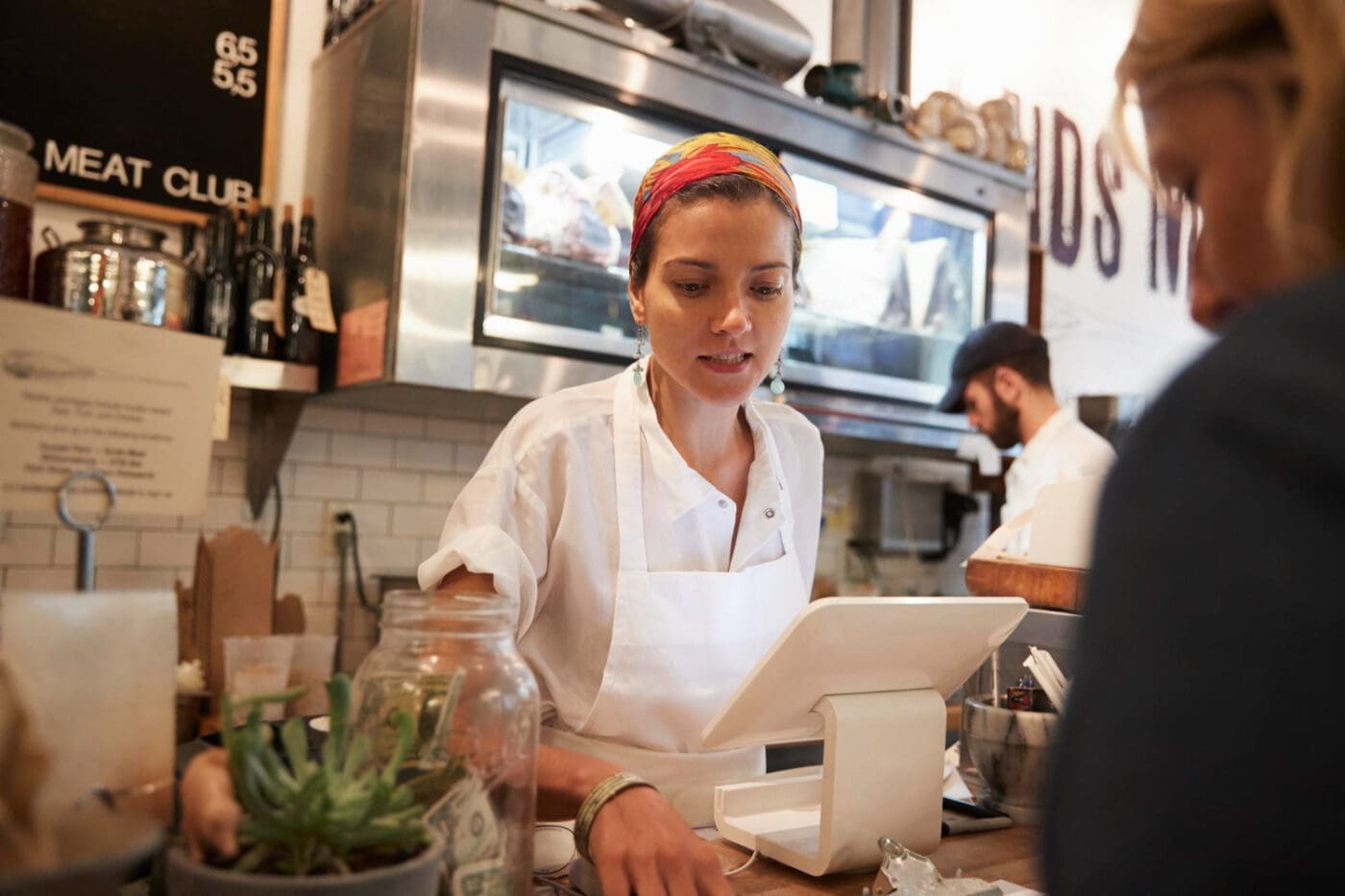 A small business owner in an apron and colorful headscarf works at the counter of her shop