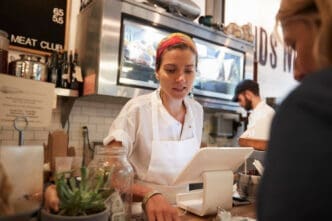 A small business owner in an apron and colorful headscarf works at the counter of her shop