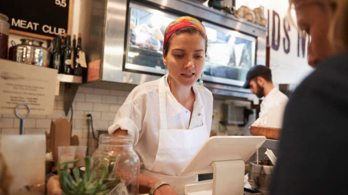 A small business owner in an apron and colorful headscarf works at the counter of her shop