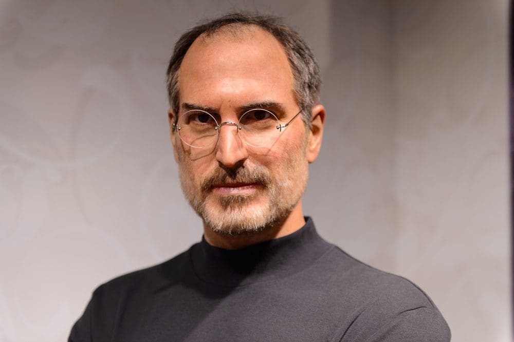 A lifelike wax figure of Steve Jobs with a beard and round glasses wearing a black turtleneck