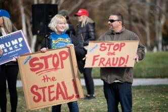 A man and a woman hold large cardboard signs that read "STOP the FRAUD!" and "STOP the STEAL!" during a political protest