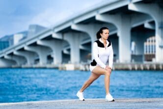 A woman in athletic wear stretches near a body of water with a large bridge in the background