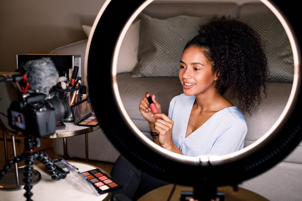 A female influencer holding a lip gloss and speaking to a camera, framed by a ring light