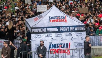 A large, diverse crowd of students and activists surrounds a promotional tent with a "Prove Me Wrong" banner