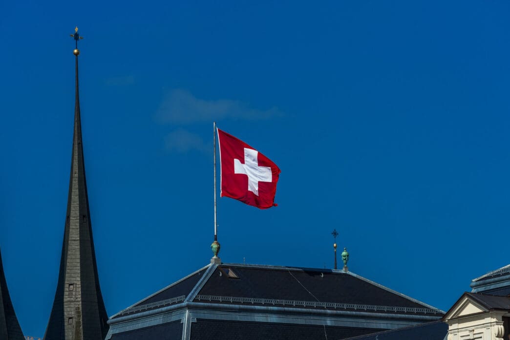 The square red flag of Switzerland with its prominent white cross waves on a flagpole atop a dark roof