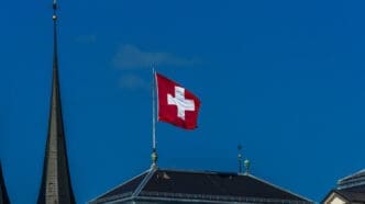 The square red flag of Switzerland with its prominent white cross waves on a flagpole atop a dark roof