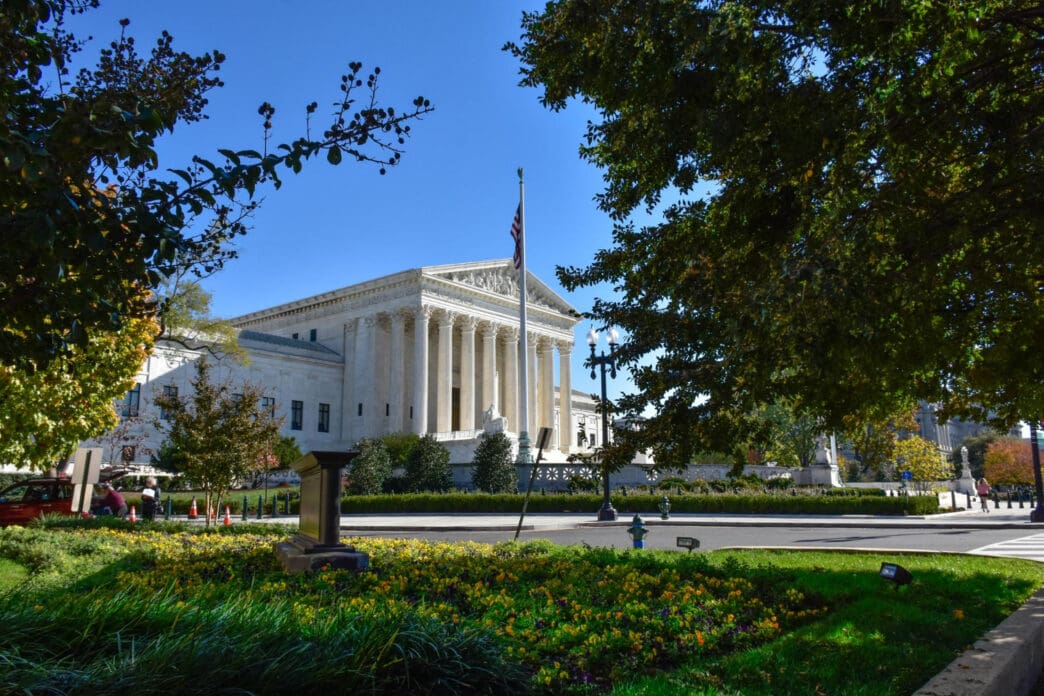 The U.S. Supreme Court building is framed by green trees and flowers on a sunny day