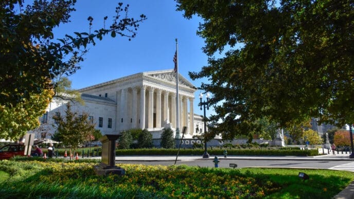 The U.S. Supreme Court building is framed by green trees and flowers on a sunny day
