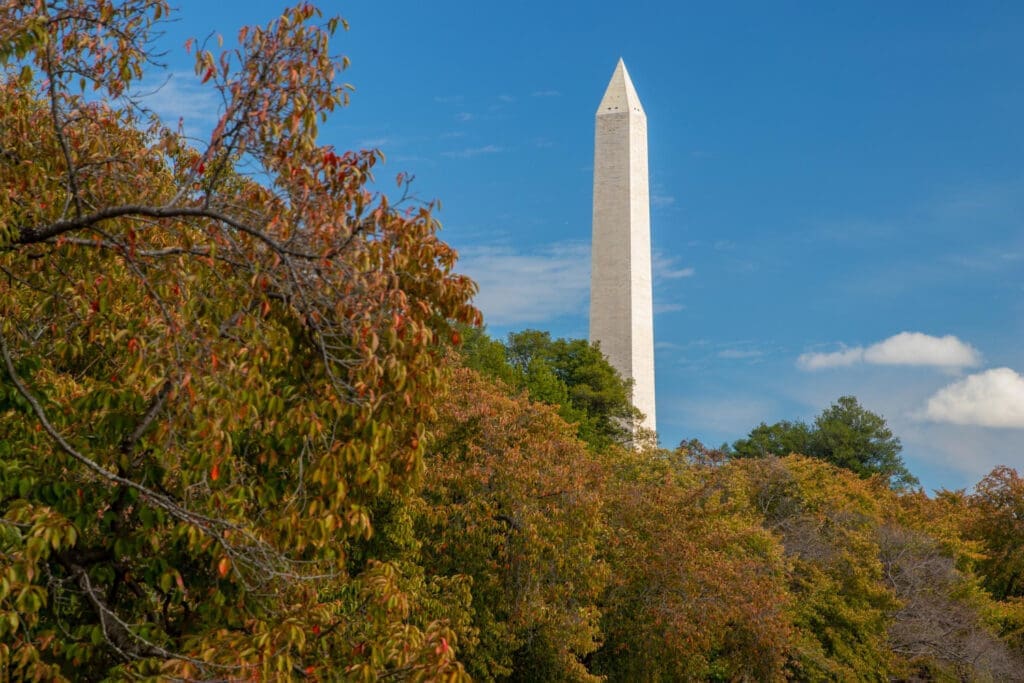 The top of the Washington Monument, a white obelisk, is visible above a hill covered in colorful autumn foliage
