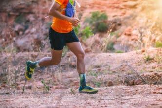 A runner wearing colorful gear runs on a dusty trail in a rocky, arid landscape
