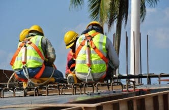 Construction workers in yellow hard hats and safety harnesses sit on a grid of rebar at a building site