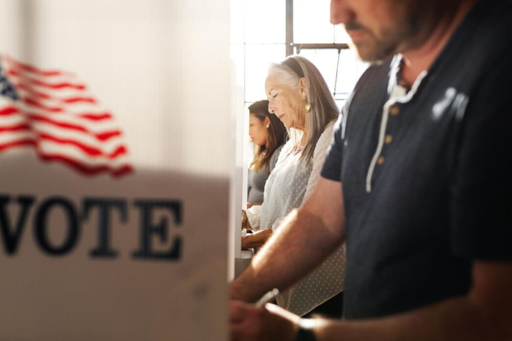 People are casting their votes inside voting booths, with a sign that reads VOTE and an American flag visible.