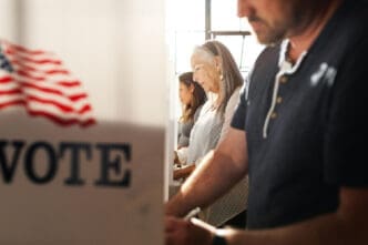 People are casting their votes inside voting booths, with a sign that reads VOTE and an American flag visible.