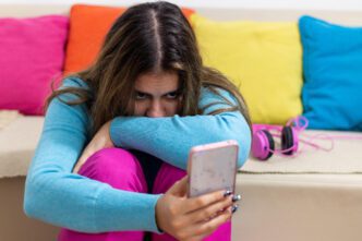 A young person looking worriedly at a smartphone, sitting on the floor with their knees hugged