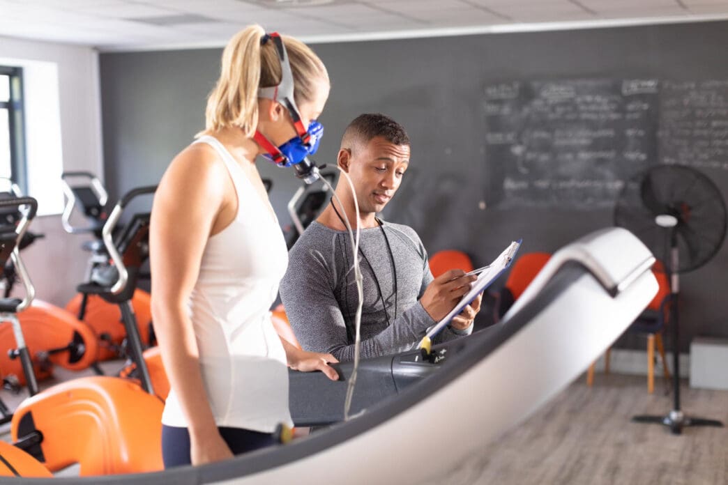 A female athlete wearing a breathing mask on a treadmill undergoes a VO2 max test while a trainer records data