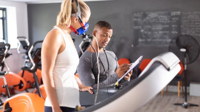 A female athlete wearing a breathing mask on a treadmill undergoes a VO2 max test while a trainer records data
