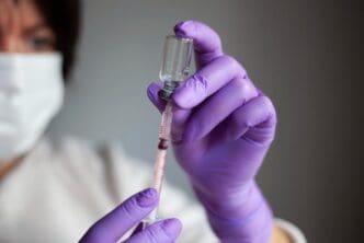 A medical professional in purple gloves draws a vaccine from a glass vial into a syringe