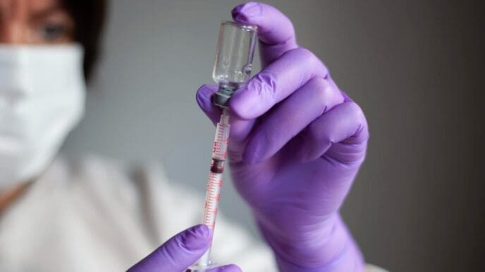 A medical professional in purple gloves draws a vaccine from a glass vial into a syringe