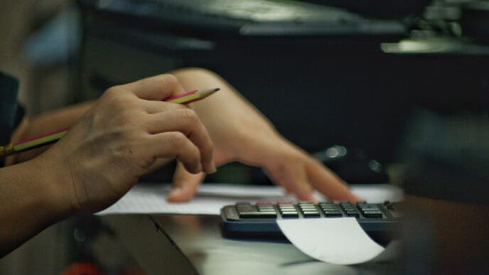 A person’s hands using a calculator and a pencil, with papers on a desk
