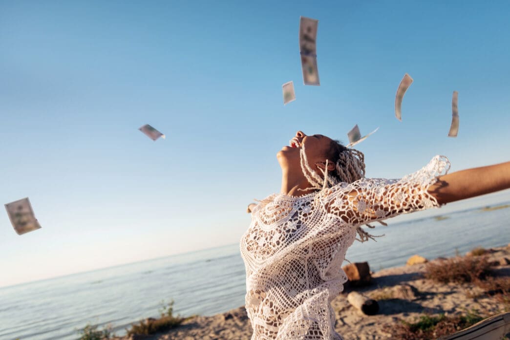 A woman throws her head back in joy as money rains down around her by the sea