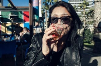Woman in a leather jacket drinking from a glass outdoors at a cafe