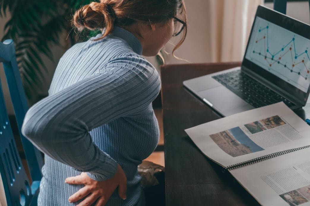 A woman experiences back pain while working at a desk with a laptop and documents