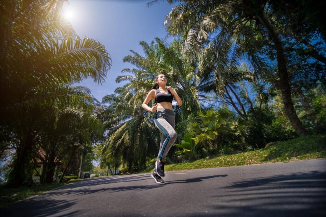 A woman in a black sports bra and gray leggings runs on a road lined with palm trees.