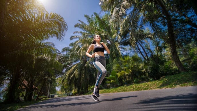 A woman in a black sports bra and gray leggings runs on a road lined with palm trees.