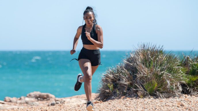 A woman running in black workout clothes on a rocky trail with the ocean in the background