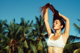 A woman with her arms raised, pulling her hair back, with palm trees and a clear sky in the background