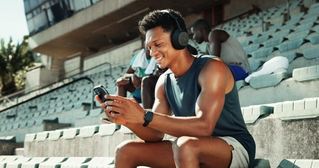 A man wearing headphones holds a phone, displaying workout metrics, likely at a stadium.