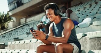 A man wearing headphones holds a phone, displaying workout metrics, likely at a stadium.