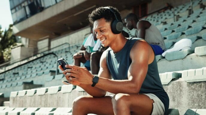 A man wearing headphones holds a phone, displaying workout metrics, likely at a stadium.
