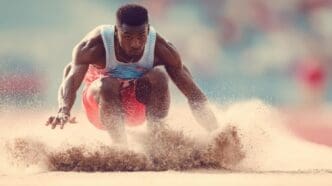 A person is mid-air, performing a long jump over a sand pit.