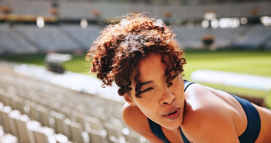 A woman in a stadium takes a break, appearing tired and sweaty after a workout.