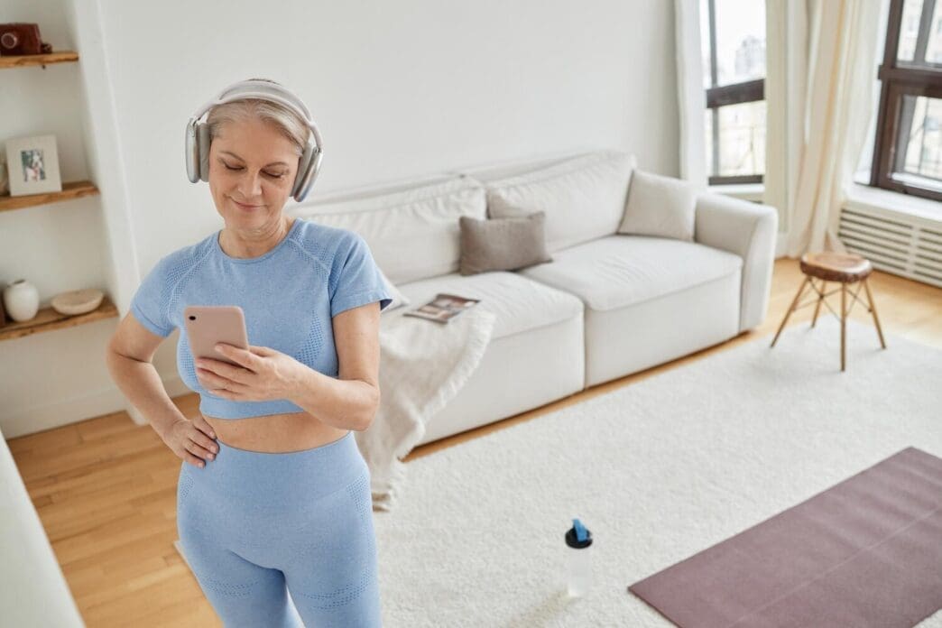 A senior Caucasian woman with headphones is exercising while using a smartphone.