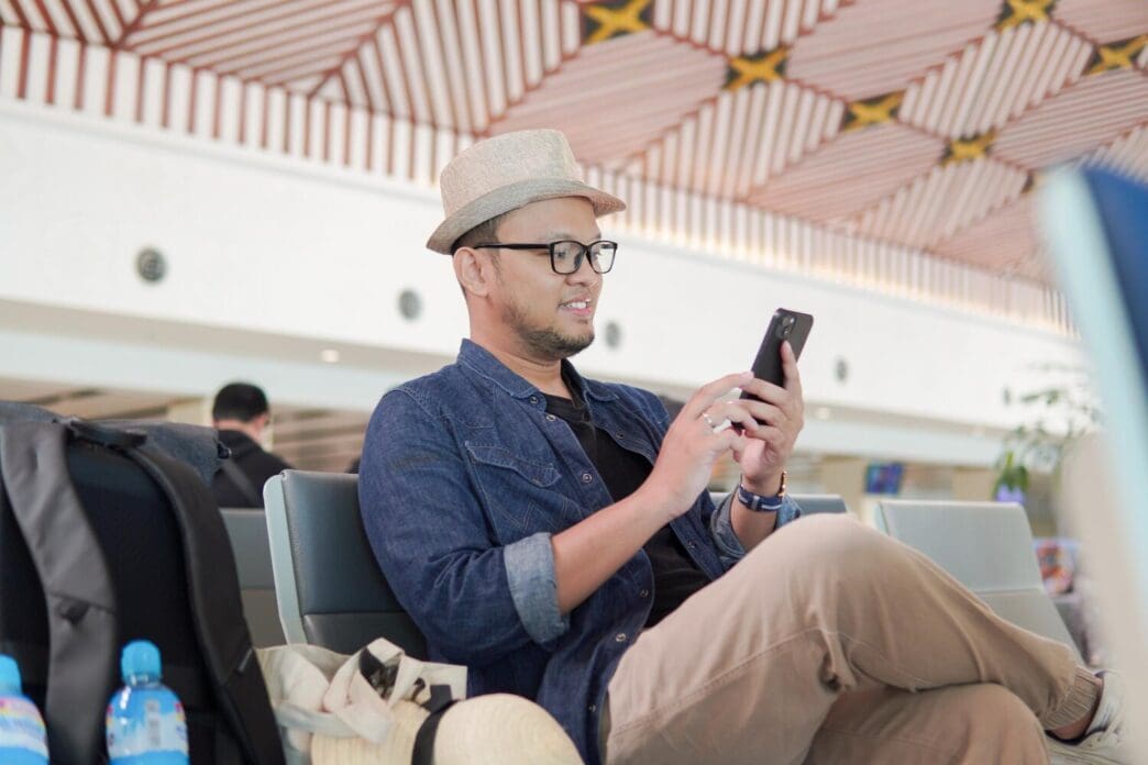 A smiling young Asian man sits on a bench in an airport lounge, holding a smartphone.