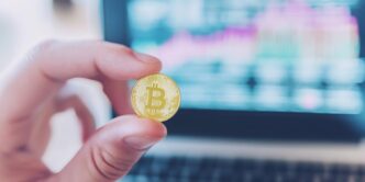 A close-up shot shows a hand holding a Bitcoin coin in front of a laptop screen displaying financial charts.