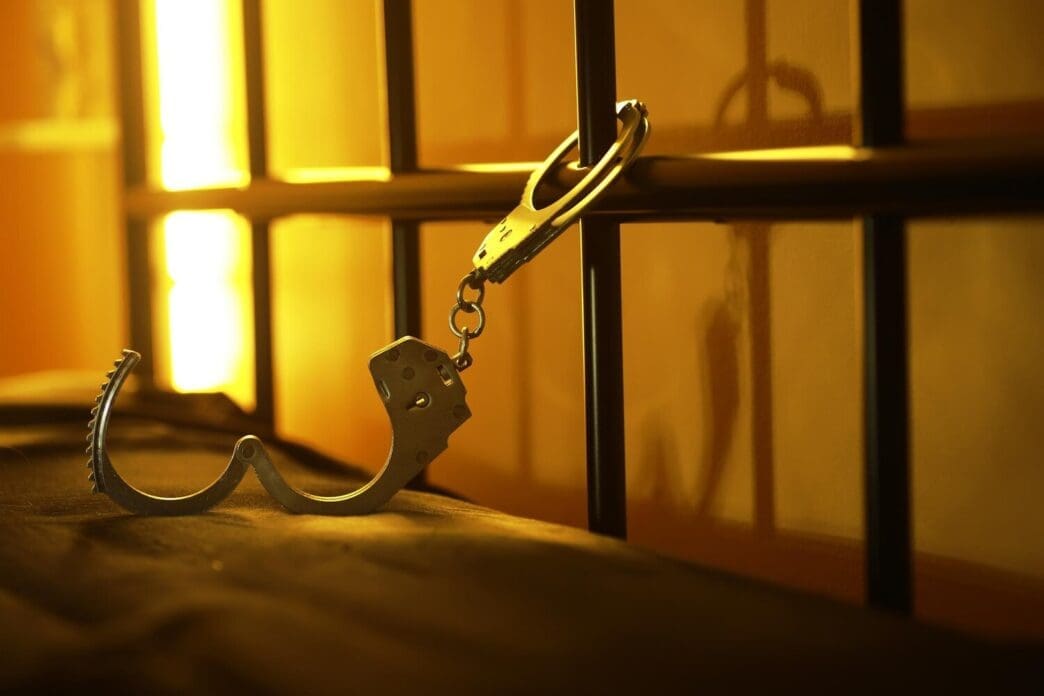 Close-up of a handcuff hanging on prison cell bars, illuminated by warm lighting.