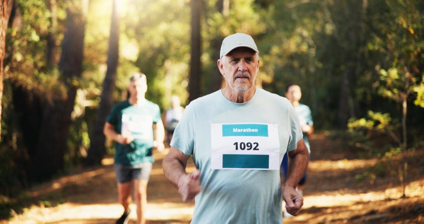 A mature man runs through a forest during a marathon, likely participating in an outdoor athletic event.