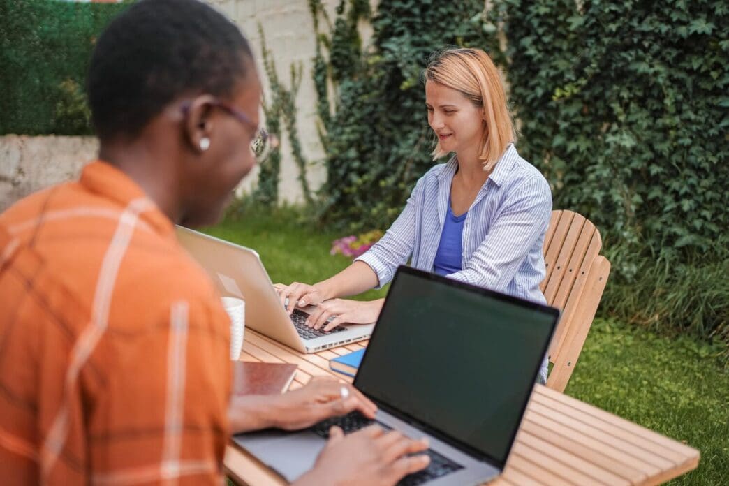 Two coworkers sit at a table in a garden, each using a laptop.