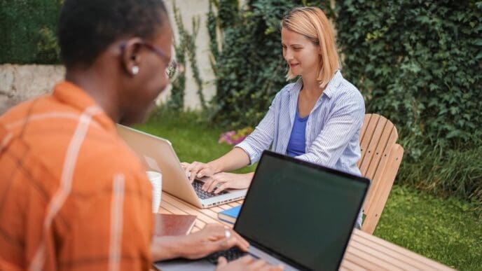 Two coworkers sit at a table in a garden, each using a laptop.