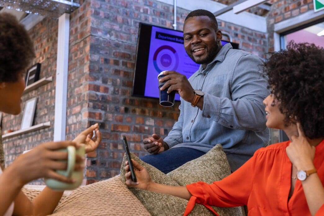 Diverse coworkers gather in an office lounge, reviewing a chart on a screen while holding travel mugs, with ample copy space.