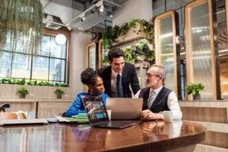 A diverse team gathers around a wooden table in a modern office, collaborating on a project displayed on a laptop screen.