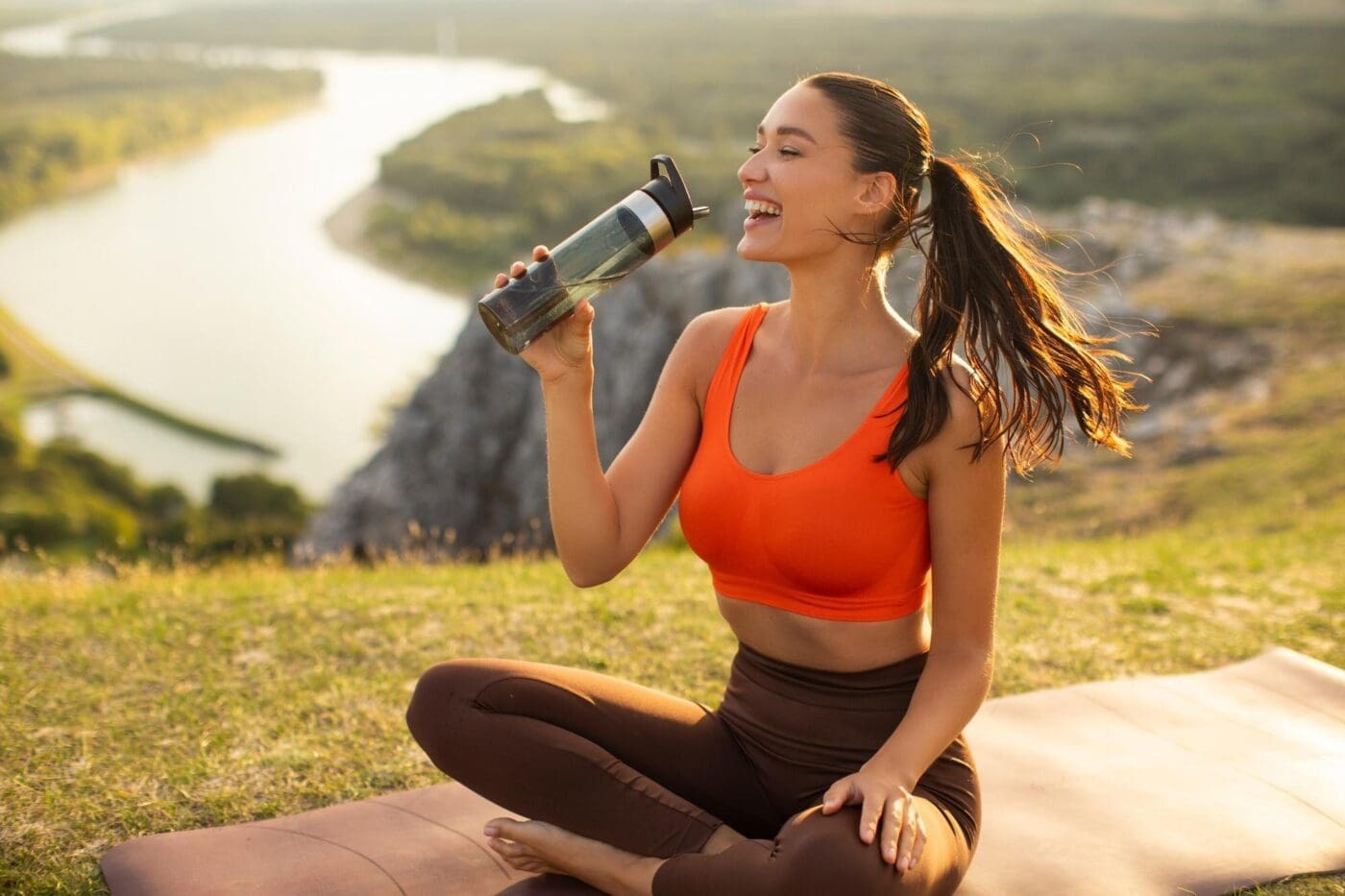 Excited woman drinks from a water bottle while sitting on a fitness mat after a yoga workout.