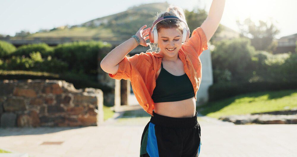 A woman in stylish streetwear dances energetically outdoors while listening to music on headphones.