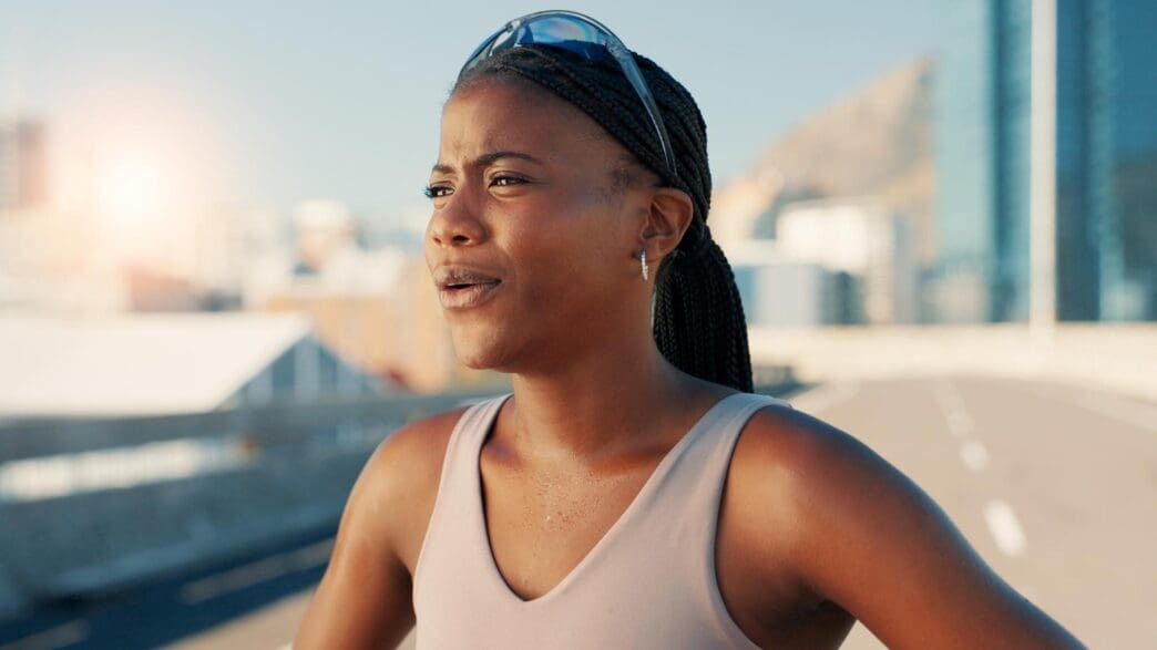 A Black woman athlete takes a break from running in an urban setting, performing a breathing exercise.