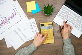 A person's hands work on a wooden desk with a laptop, calculator, and papers related to financial analysis.