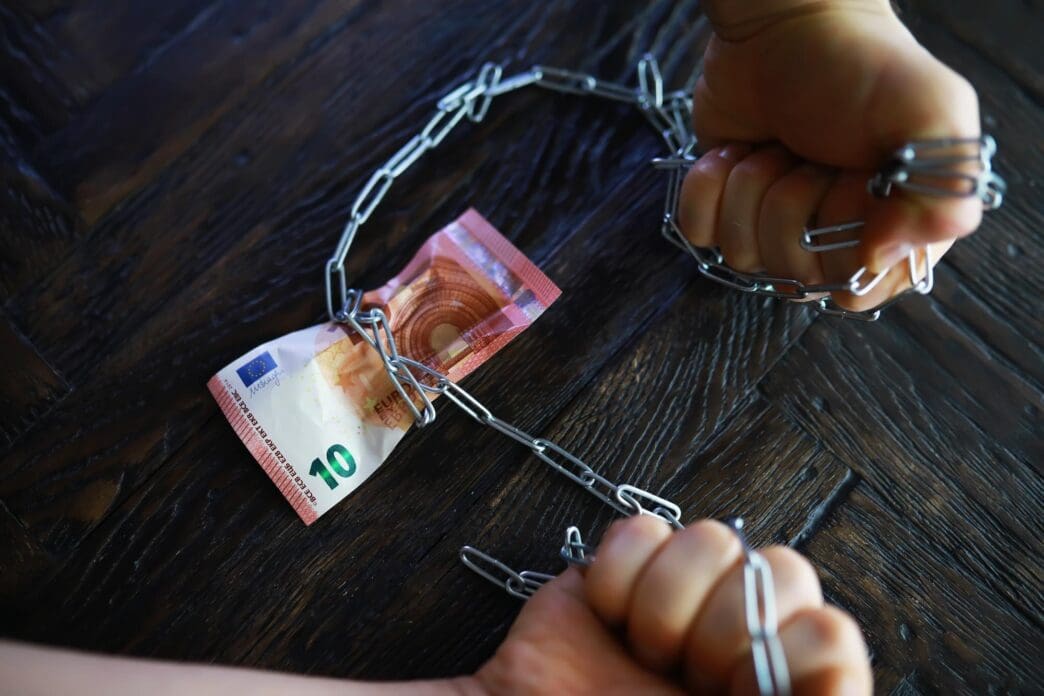 Hands chained with metal links struggle to free a banknote.